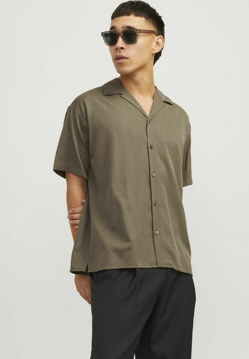 Vyras vilkintis alyvuogių žalios spalvos trumpomis rankovėmis marškinius su sagomis ir juodomis kelnėmis, žiūri į šoną, su tamsiais akinių nuo saulės ir viena ranka už nugaros.