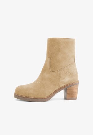 Bottine en suède beige avec un talon carré, un bout rond et un dessus lisse. Présente peu de coutures et un logo discret.