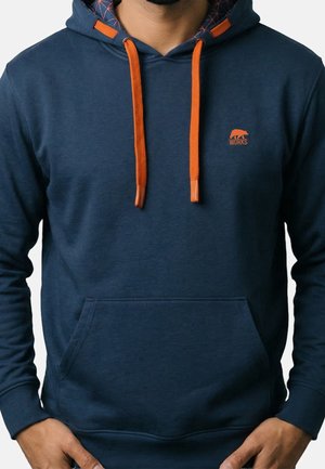 Homme portant un sweat à capuche bleu marine avec des cordons orange et un petit logo ours orange avec le mot « WORKS » sur la poitrine.