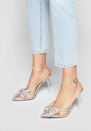 Fødder iført lyseblå jeans, der er bukket op over anklen, og gennemsigtige høje hæle med rhinsten sløjfer og ankelremme.