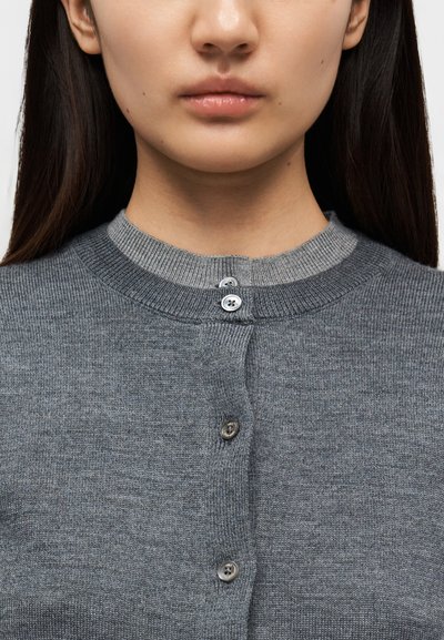 Γκρι πλεκτή ζακέτα με στρογγυλή λαιμόκοψη και δύο κουμπιά μπροστά. Διαθέτει αντίθετη υφή πλέξης κατά μήκος του γιακά.