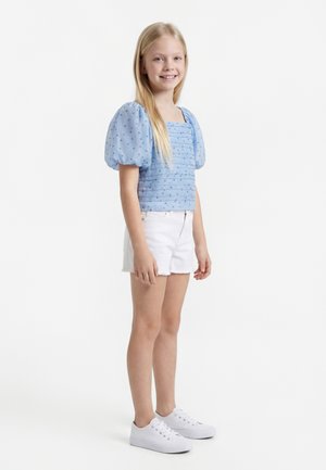 Ragazza in piedi e sorridente, indossa una maglietta blu con maniche a sbuffo a pois, pantaloncini bianchi e sneakers bianche su sfondo bianco.