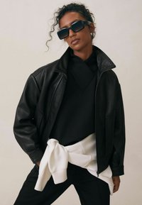 Czarna skórzana kurtka o teksturowanym wykończeniu, noszona na czarnym topie, z białą bluzą związana w talii. Modelka w dużych okularach przeciwsłonecznych.