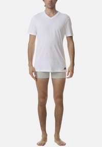 Biała koszulka z krótkim rękawem i dekoltem w serek, połączona z jasnoszarymi szortami, z logo Adidas na dolnym lewym boku. Ubrane przez modela stojącego boso.