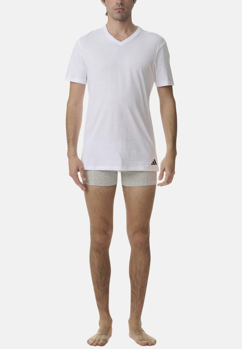 Biała koszulka z krótkim rękawem i dekoltem w serek, połączona z jasnoszarymi szortami, z logo Adidas na dolnym lewym boku. Ubrane przez modela stojącego boso.