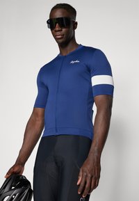 Navy-Fahrradtrikot mit kurzen Ärmeln, weißer Streifendetail an den Ärmeln, Front-Reißverschluss, kombiniert mit schwarzen gepolsterten Shorts und einem schwarzen Helm.