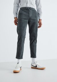 Graue Cordhose mit verstellbarem Bund, schmalem Schnitt und gerippter Textur, kombiniert mit weißen Turnschuhen und Socken in Crew-Länge.