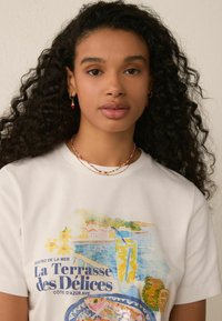 White cotton t-shirt featuring a colorful seaside graphic print with text. Accessories include a beaded necklace and earrings. Curly hair visible.