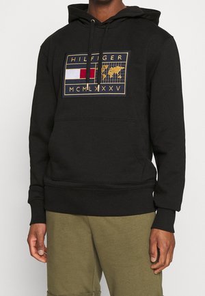 Czarna bluza z kapturem z przednią kieszenią, złotą naszywką z czerwono-biało-niebieską flagą, mapą świata, napisem "HILFIGER" i cyframi rzymskimi MCMLXXXV.