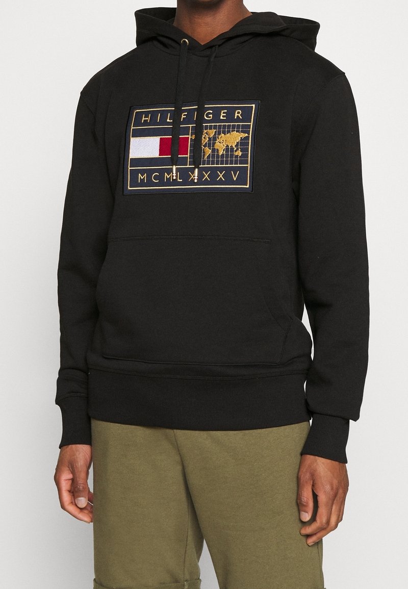 Sweat à capuche noir avec poche kangourou avant, écusson bordé d'or affichant un drapeau rouge, blanc et bleu, carte du monde, "HILFIGER" et chiffres romains MCMLXXXV.
