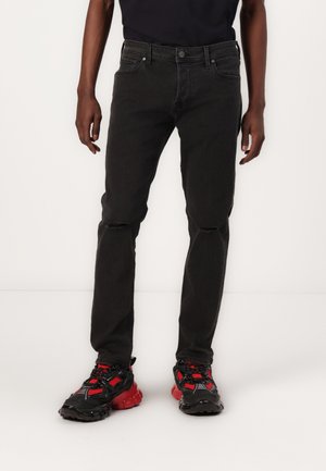 Black denim jeans with a slim fit, featuring slight distressing at the knees, five pocket design, and subtle fading. Paired with black and red sneakers.