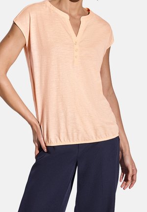 Kobieta ubrana w jasnoróżową koszulkę henley z krótkim rękawem i guzikami oraz ciemnoniebieskie luźne spodnie, pozuje z jedną ręką na biodrze.