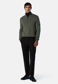 Pullover verde a maglia con zip a quarti e polsini a coste, abbinato a pantaloni neri e sneakers nere. Nessun logo o motivo visibile.