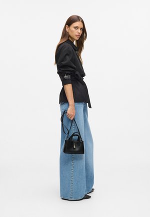 Frau in schwarzer taillierter Jacke und blauen weiten Jeans, die eine kleine schwarze Handtasche hält, vor einem weißen Hintergrund stehend.