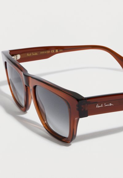Gafas de sol translúcidas marrones con lentes gris oscuro. Diseño angular, con un frente negro contrastante y el logo en relieve de "Paul Smith" en las patillas.