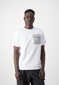 Hvid bomuld t-shirt med en grå stoflomme. Korte ærmer, rund hals og diskret sidesøm label detalje. Glat tekstur.