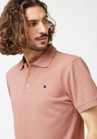 Polo in rosa polveroso con un tessuto texturizzato. Presenta un colletto, tre bottoni e un piccolo logo di un albero blu navy sul lato sinistro del petto.