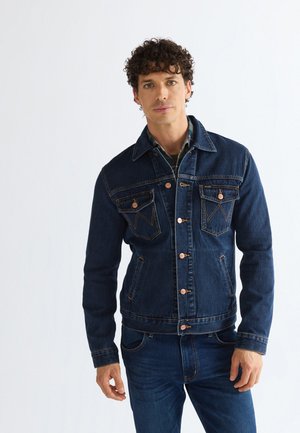 Homme aux cheveux bouclés portant une veste en jean foncé et un jean, debout devant un fond blanc uni.