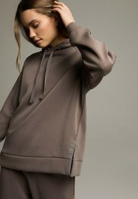 Bluza z kapturem w stonowanym brązie, charakteryzująca się luźnym krojem, teksturowanym materiałem oraz bocznym zamkiem dla zwiększenia funkcjonalności.