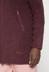 Vínová fleecová bunda s texturovaným povrchem, celopropínacím zipem a bočními kapsami. Má značkové logo poblíž lemu.