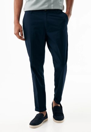 Ein Mann trägt marineblaue, maßgeschneiderte Hosen mit umgeschlagenen Säumen und schwarze Loafers mit weißer Sohle, er steht mit den Händen seitlich am Körper.