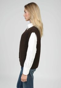 Veste en tricot marron avec un motif texturé, portée sur une chemise blanche, associée à un jean bleu, présentée de profil.