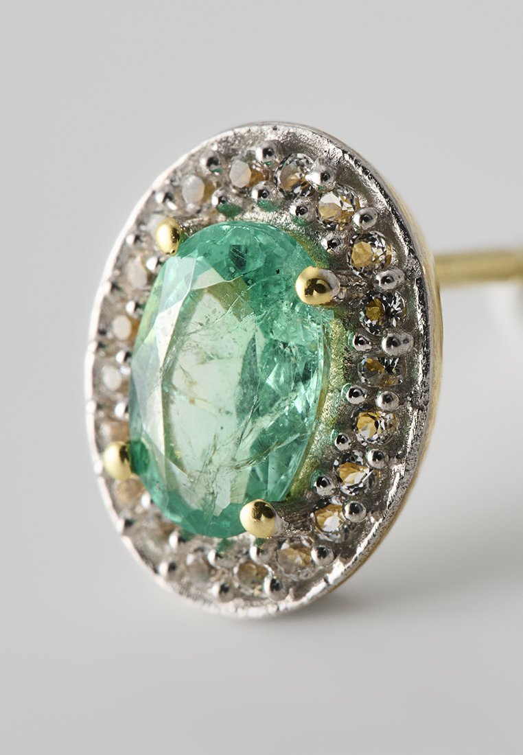 Boucle d'oreille ovale en argent avec une grande pierre précieuse verte entourée de petites pierres brillantes, mise en valeur par des accents dorés.