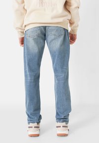 Hellblaue Jeans mit geradem Schnitt, ausgestattet mit zwei Gesäßtaschen und subtilen Auswaschungen. Getragen mit einem beigen Sweatshirt und Sneakers.