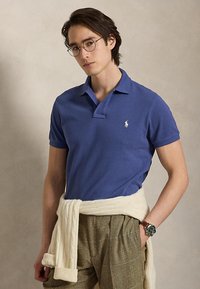 Junger Mann mit blauem Poloshirt, karierten Hosen, cremefarbenem Pullover um die Taille gebunden, runden Brillen und Armbanduhr, steht vor schlichtem Hintergrund.