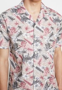 Camisa de manga corta en beige claro con un estampado floral en rosa, gris y blanco. Cuenta con frente de botones y cuello. Material de algodón.