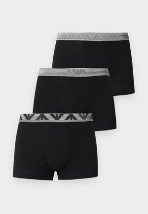 Mladý muž s krátkymi tmavými vlasmi nosiace čierne priliehavé boxery so sivým vzorovaným pásom, stojaci bosý na jednolennom bielom pozadí.