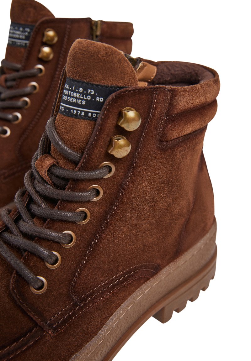 Botas de tobillo de ante marrón con punta redonda, cordones negros, herrajes de latón, cuello acolchado y suela de goma texturizada. Etiqueta de la marca visible.
