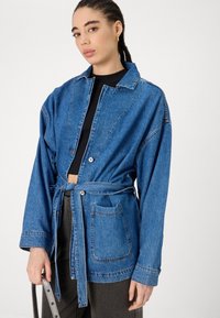 Denim jakna s pasom u struku, dugim rukavima i džepom na zakrpu. Karakterizira je svijetloplava boja i opušteni kroj s vidljivim šavovima.