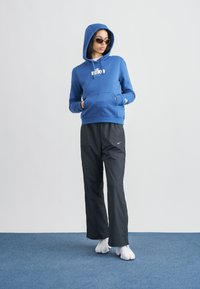 Μπλε φούτερ με κουκούλα και μπροστινή τσέπη, με λευκό λογότυπο, σε συνδυασμό με μαύρο φαρδύ παντελόνι, φορεμένο με λευκά παπούτσια και γυαλιά ηλίου.