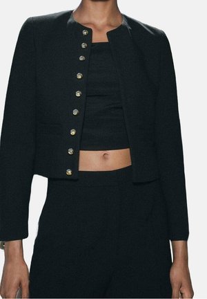Femme portant une veste courte noire à boutons dorés par-dessus un crop top noir et un pantalon noir taille haute, laissant voir la région du ventre.