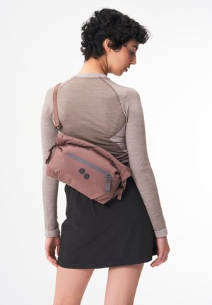 Rosa Umhängetasche aus weichem Material, mit einer grauen Reißverschlusstasche und zwei schwarzen runden Akzenten. Verstellbarer Trageriemen ist enthalten.