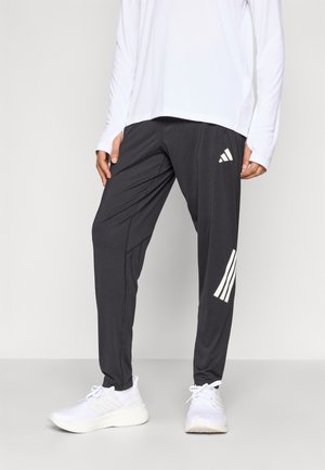 Personne portant un pantalon de survêtement Adidas noir avec des bandes blanches et des baskets blanches, debout devant un fond clair uni.