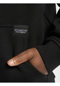 Fekete kapucnis pulóver kenguru zsebbel és bordás mandzsettákkal. A zseb felett egy fehér ROC-A-WEAR címke látható. Puha szövet textúra.