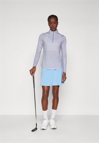 Langermet golftrøye med blå og hvite mønstre, med glidelås i kragen. Kombinert med en lyseblå skjørt og hvite joggesko.