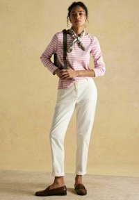 Kobieta stoi, ubrana w różowo-białą w paski koszulę z długim rękawem, białe spodnie, brązowe mokasyny, kwiatowy szalik na szyi i nosi torbę przewieszoną przez jedno ramię.