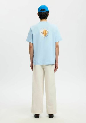 Personne portant une casquette bleue, un t-shirt bleu clair avec un patch circulaire représentant un croissant dans le dos, un pantalon crème à jambes larges, vue de dos.