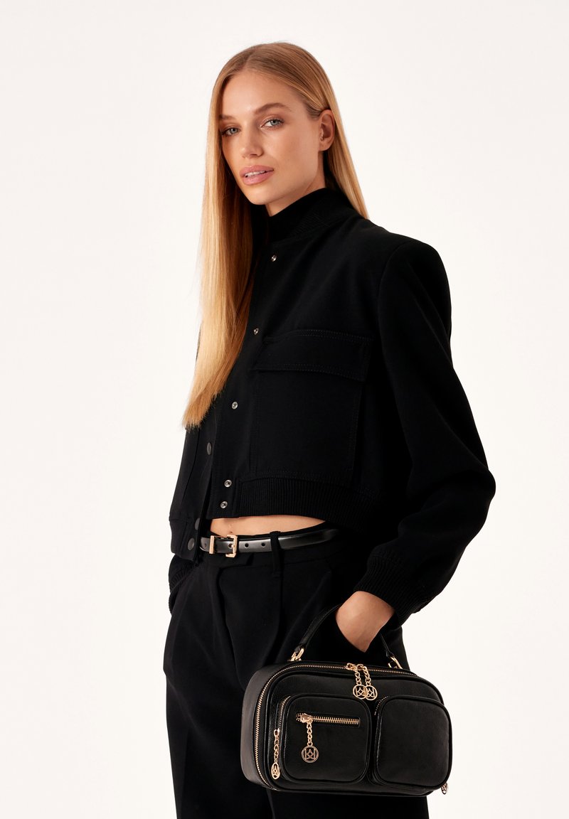 Schwarze, verkürzte Strickjacke mit Druckknöpfen und Taschen, kombiniert mit hochtaillierten schwarzen Hosen. Hält eine strukturierte schwarze Handtasche mit goldenen Akzenten.