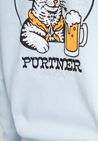 Chat portant une écharpe orange assis à côté d'une chope de bière mousseuse avec le texte "PURTNER Volcom Stone" sur un tissu bleu clair.
