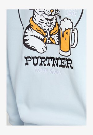 Kat met een oranje sjaal zittend naast een schuimend glas bier met de tekst "PURTNER Volcom Stone" op lichtblauwe stof.