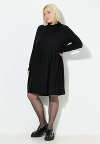Sort langærmet kjole med høj hals og indsamlet talje. Den er lavet af blødt materiale og har en svajende nederdel samt en glat tekstur.