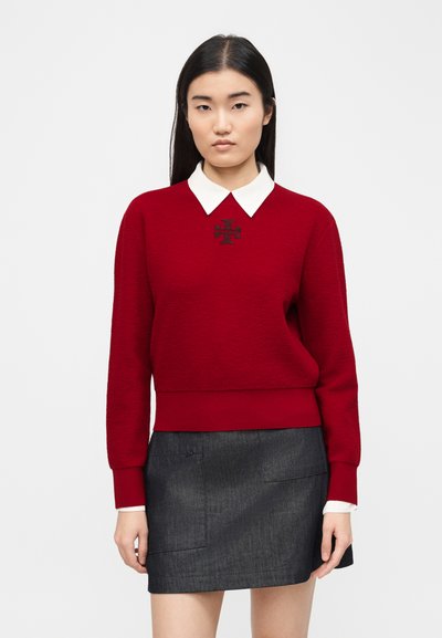Femme portant un pull rouge avec un col blanc et un logo noir, associé à une mini-jupe gris foncé, debout devant un fond uni.