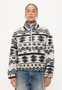 Fleece mikina s polovičním zipem, která má geometrický vzor v černé a bílé barvě, světle modrý zip a měkký, strukturovaný povrch.