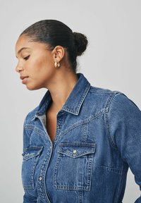 Modrá denimová košela s dvoma prednými vreckami, patentnými zapínaniami a golierom. Hladká textura, priliehavý strih a viditeľné švy.