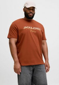 Pruun puuvillane T-särk, millel on valge ja kuldne "JACK & JONES" logo, kombineeritud tumehallide teksashortsidega. Modelli peas on heledat värvi müts.