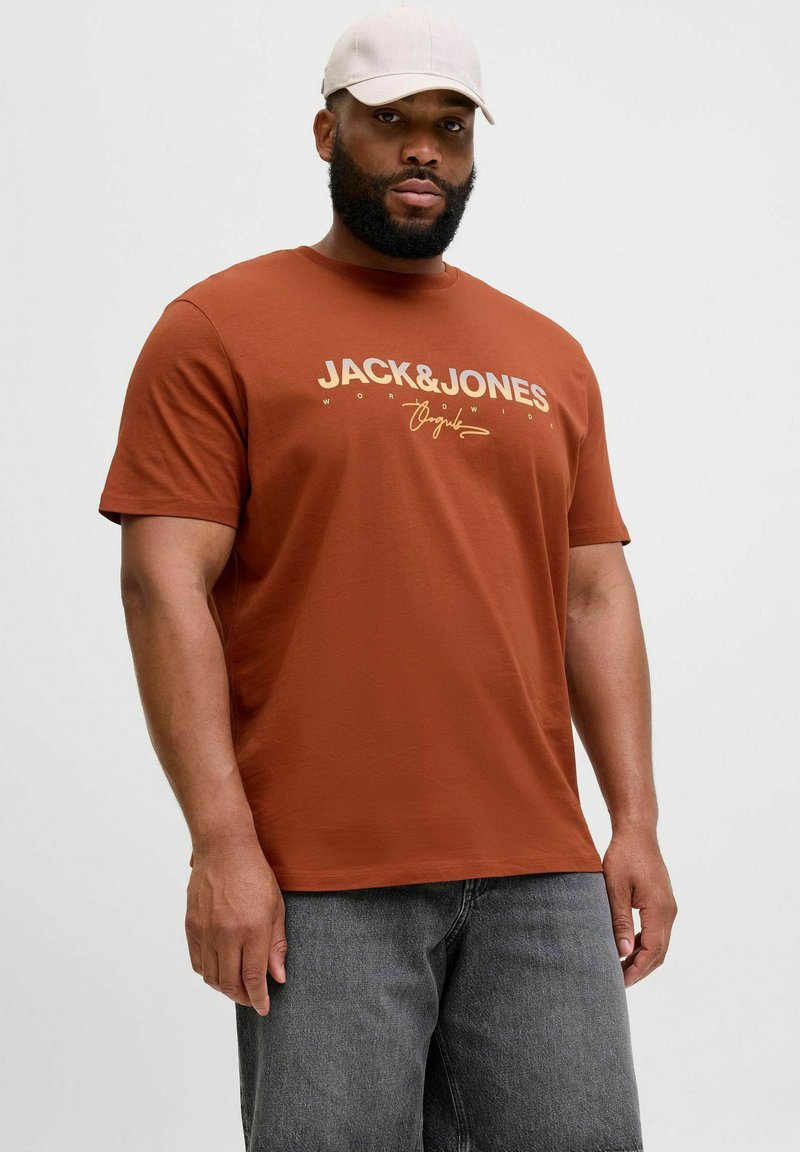 Pruun puuvillane T-särk, millel on valge ja kuldne "JACK & JONES" logo, kombineeritud tumehallide teksashortsidega. Modelli peas on heledat värvi müts.